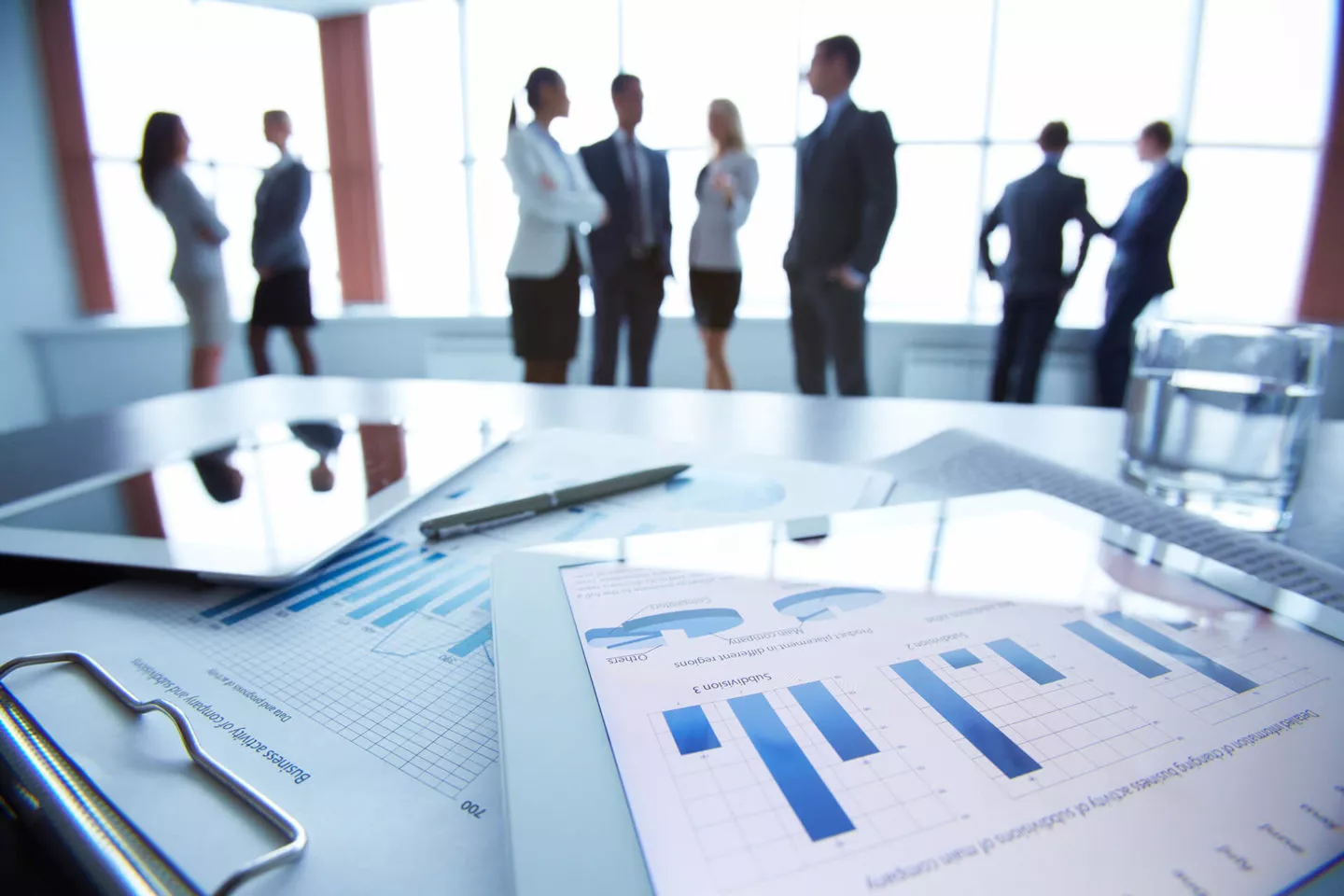 Paper reports on a table with groups of business leaders standing in the background talking with each other.