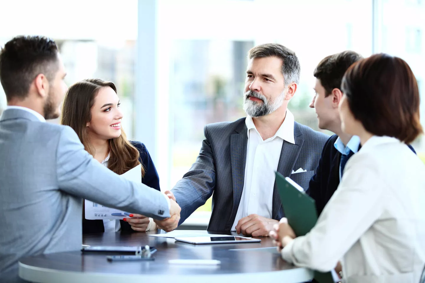 A group of five business leaders wearing professional attire stand around a tall round table. Two men shake hands.
