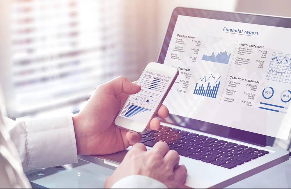Man comparing financial report data on laptop and mobile phone.