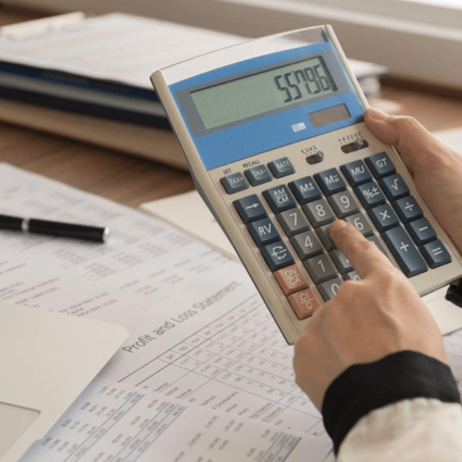 Hands using a calculator for bookkeeping. A profit and loss statement and other financial documents are scattered on a desk.