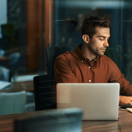 A male professional in a brown button down shirt sitting at a desk with his laptop.