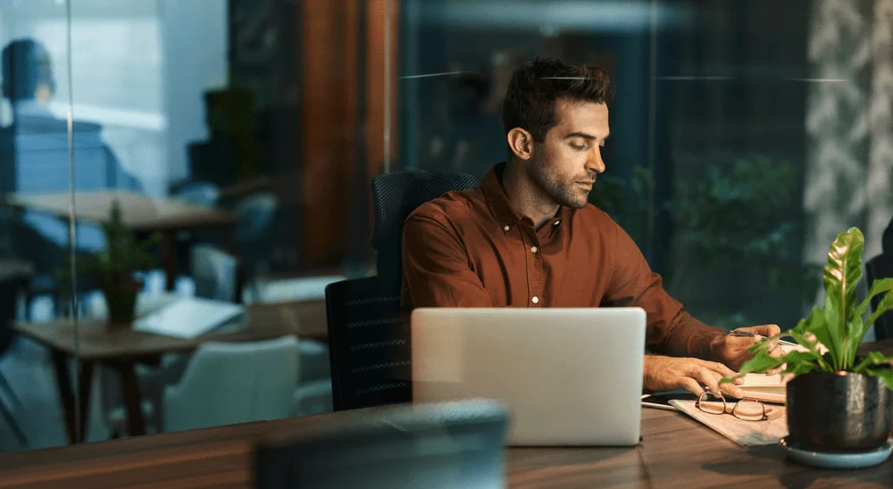 A male professional in a brown button down shirt sitting at a desk with his laptop.
