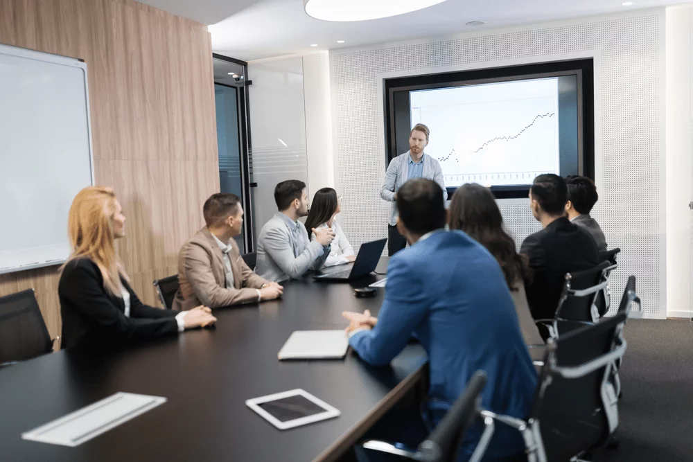 A boardroom meeting of business professionals with one man upfront presenting data from a graph.