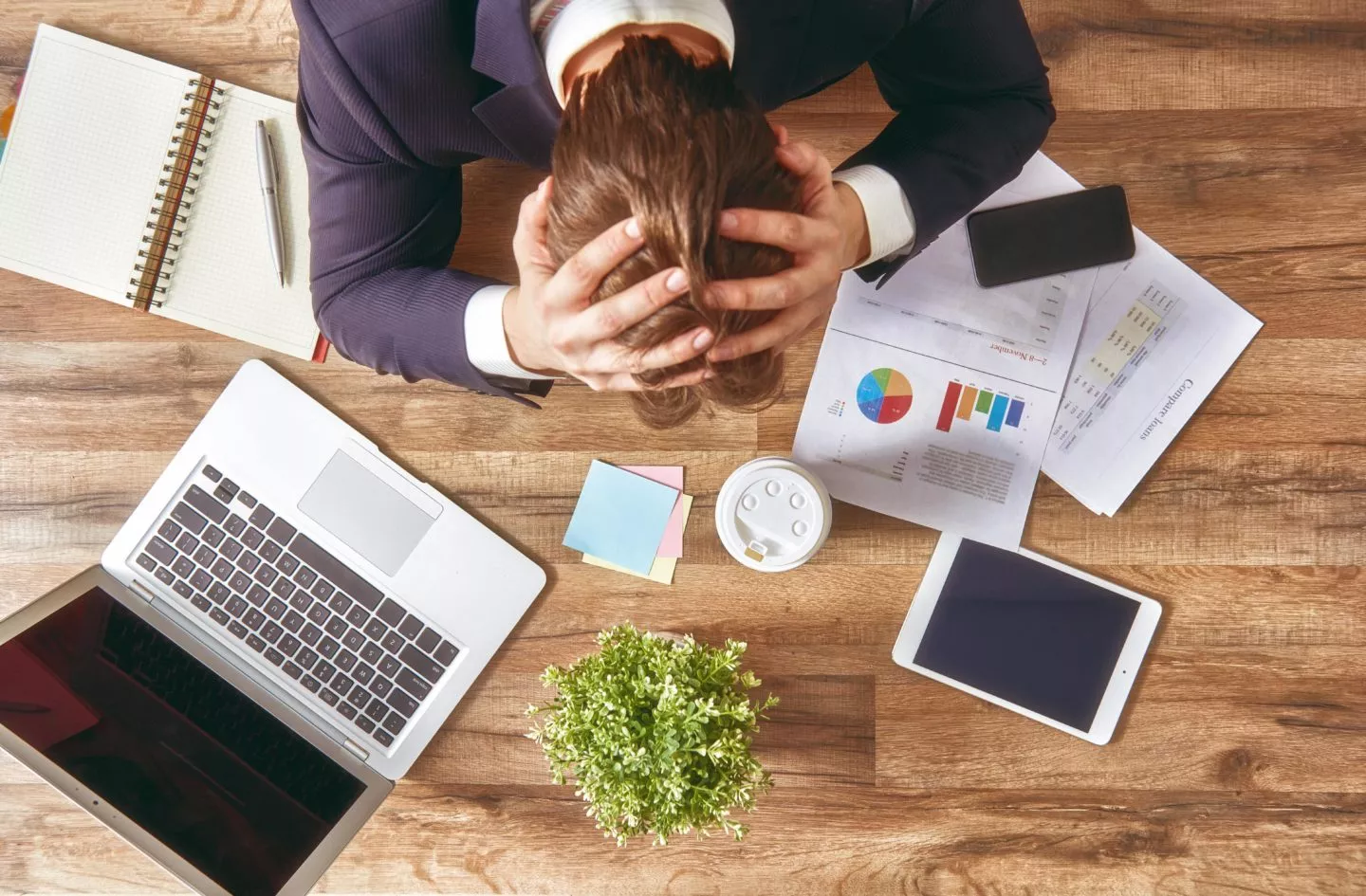 A stressed out man holding his head while looking down at a table with financial documents and a laptop open.