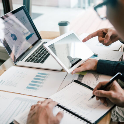 Two people working on a laptop and tablet while examining financial documents.
